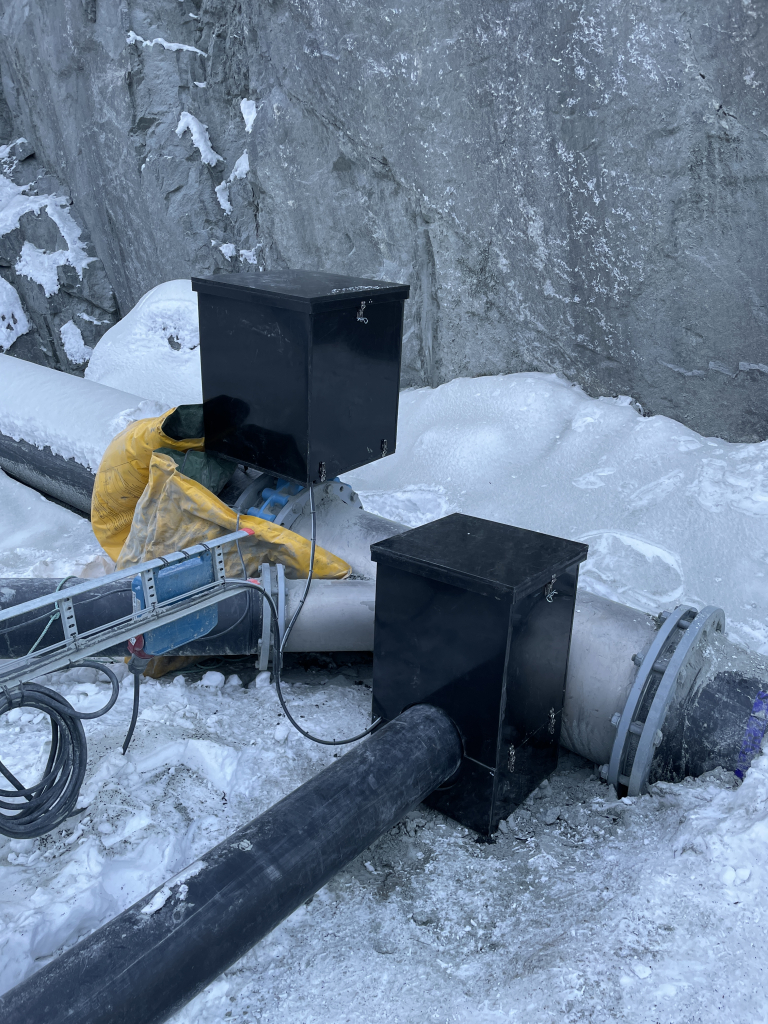 Industrielle rør og svarte kontrollbokser montert i et snødekt, steinete terreng